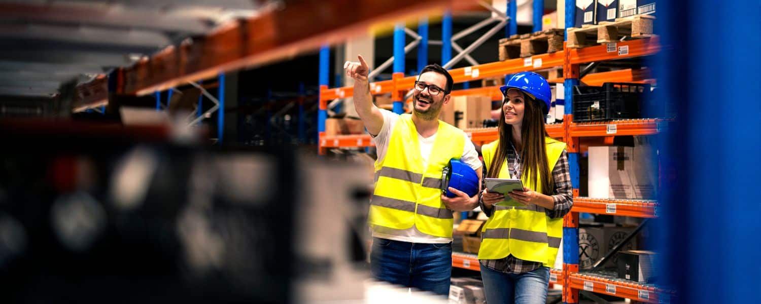 Two warehouse managers view product shelving whilst comparing information on digtial tablet. Image used to represent blog post titled "Choosing the right catalogue automation tool for your business"
