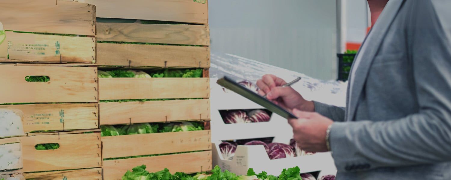 Close up image of a stacked wooden crates of fresh vegetables, with an employee taking inventory details via a digital tablet. Image used to represent a blog titled "EDI in the Supply Chain. Exploring the challenges faced by food and drinks brands when supplying large retailers"