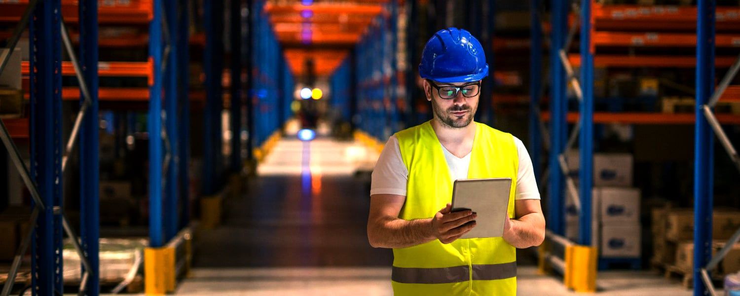 Warehouse Manager wearing hardhat and high vis jacket reviews digital catalogues on an electronic tablet whilst stood on the warehouse floor