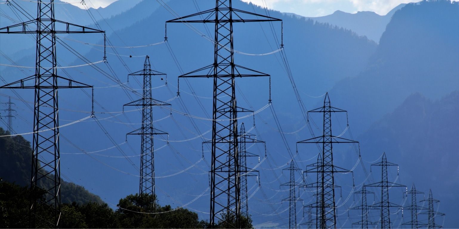Blue hued landscape photo of a series of power lines into the distance, with tree covered mountains in the background. Used to represent the concept of procurement technology connecting organisations to a network of suppliers.