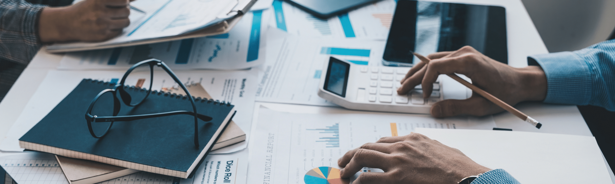 Blog feature image showing a close up of a messy desk with scattered papers; one pair of hands holds a clipboard and another pair of hands use a calculator. It represents the AP and AR teams working hard to fulfil the invoicing process.