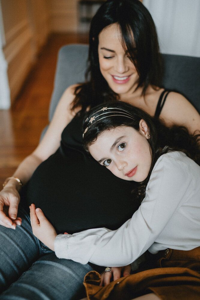 Girl holds pregnant mother's belly and looks at photographer
