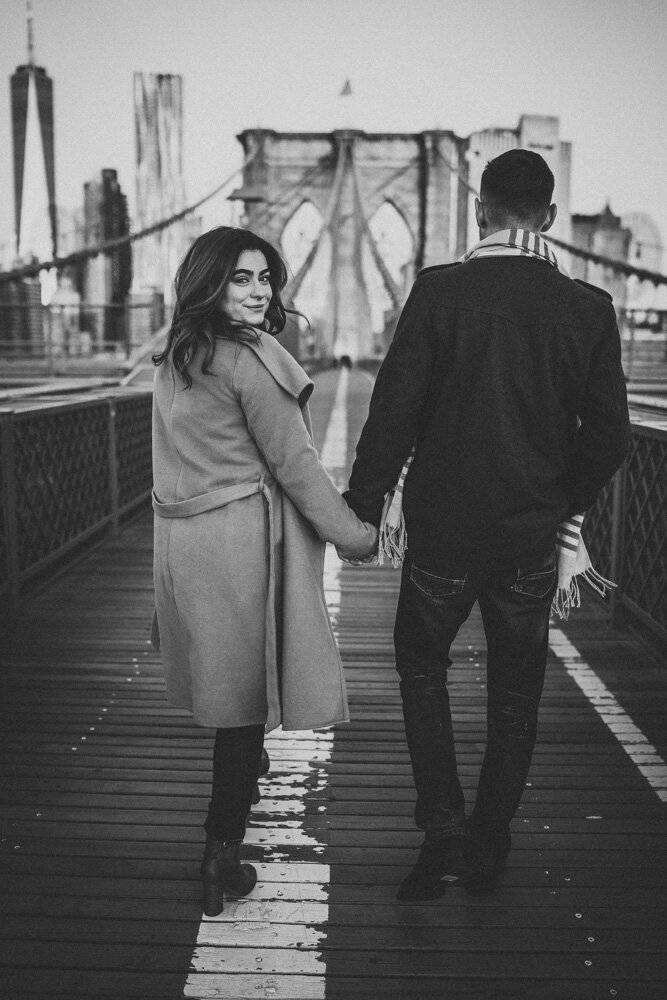 Woman holds boyfriend's hand on brooklyn bridge and glances back at photographer