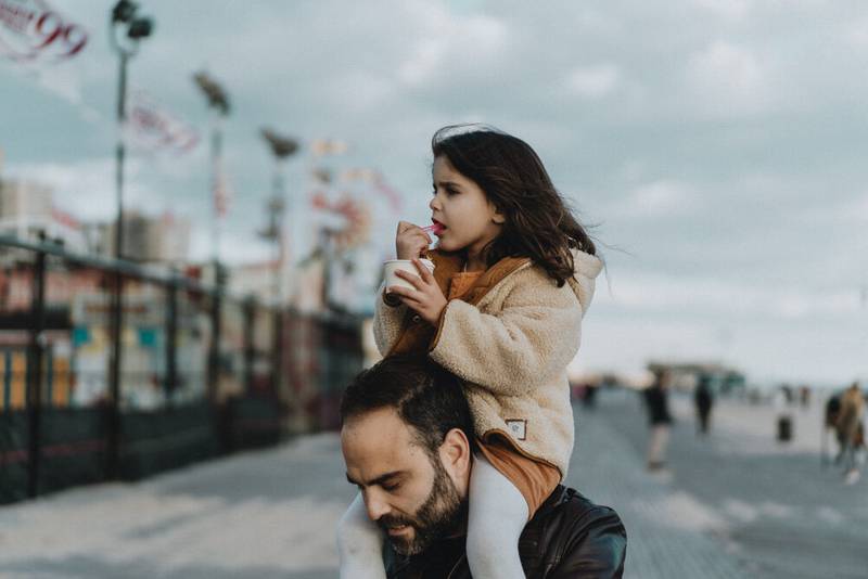 Young girl eats ice cream on dad's shoulders during family session at coney island
