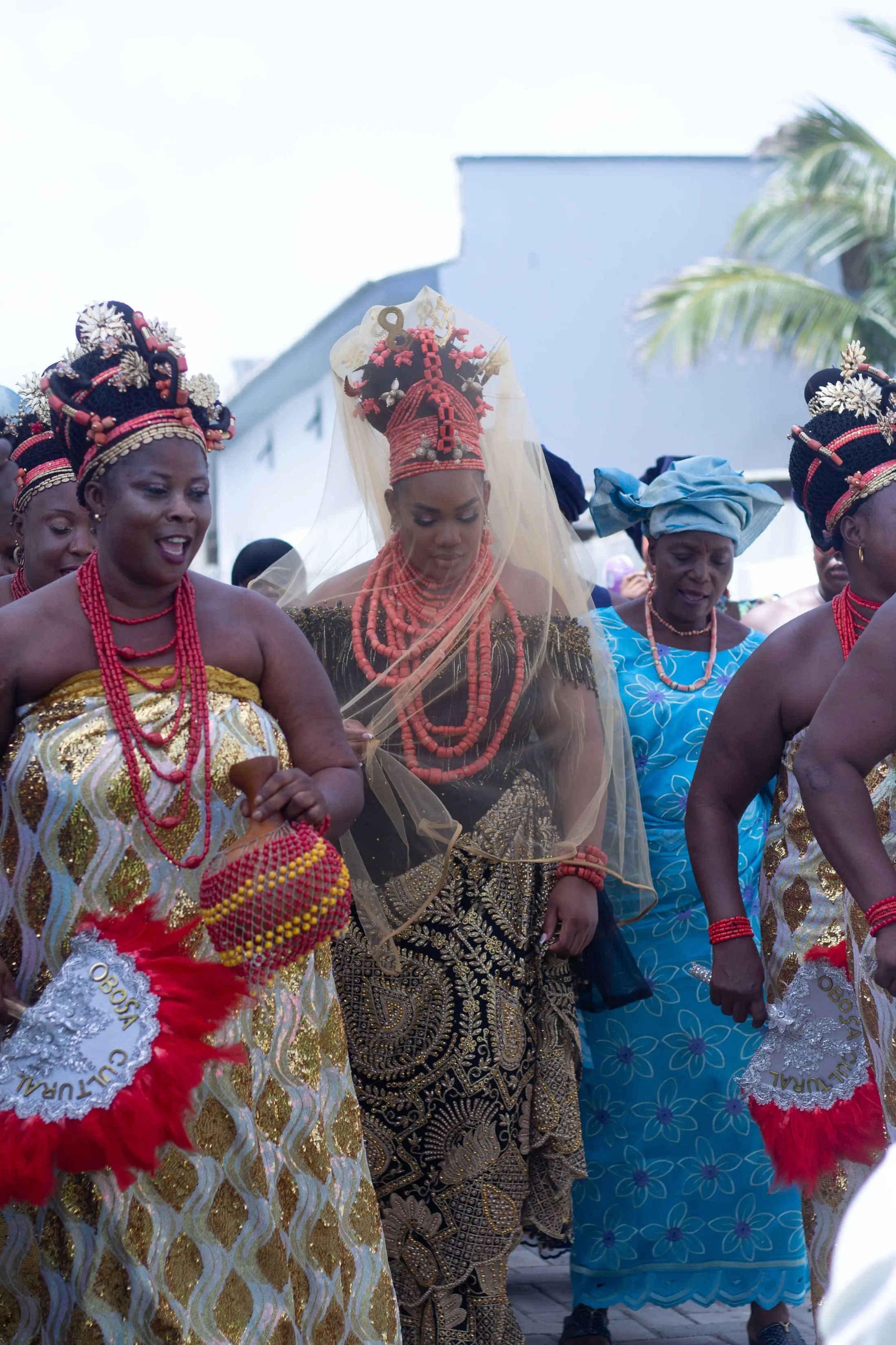 Women in coral beads and gold wrappers dancing in with red feather fans