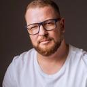 Homem branco de cabelo preto e barba bem feita, usa óculos e camiseta branca. 