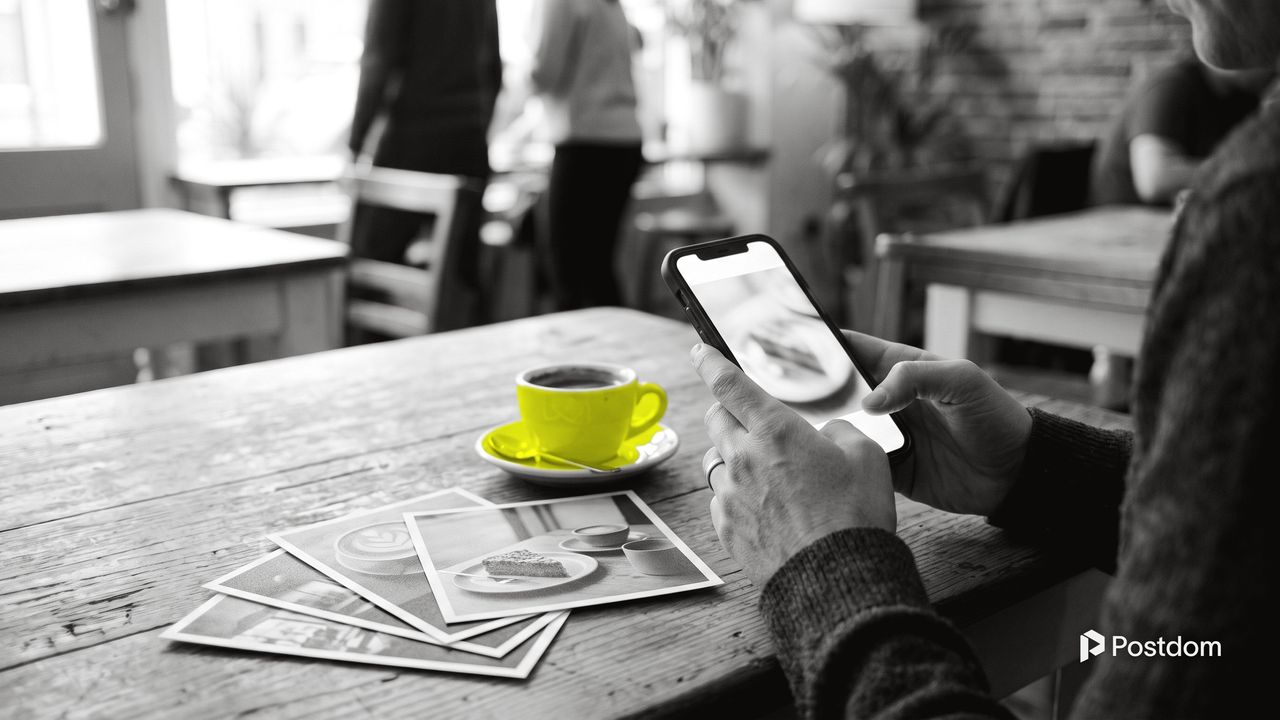Best AI social media content generator: a café owner checks a phone next to printed photos of coffee and cake and a yellow espresso cup