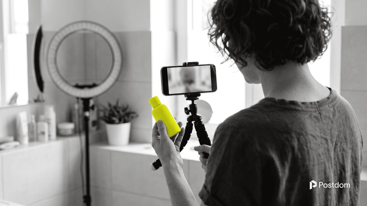 Best AI UGC tools: a woman seen from behind holds a yellow skincare bottle up to a phone on a small tripod in a bright bathroom