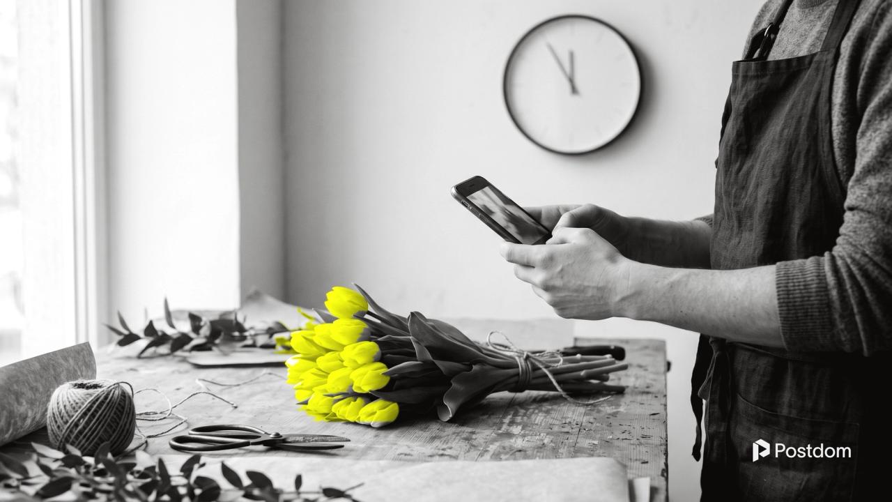 Best time to post on Instagram: a florist photographs a bunch of yellow tulips on a wooden workbench with a phone, a wall clock just before noon behind