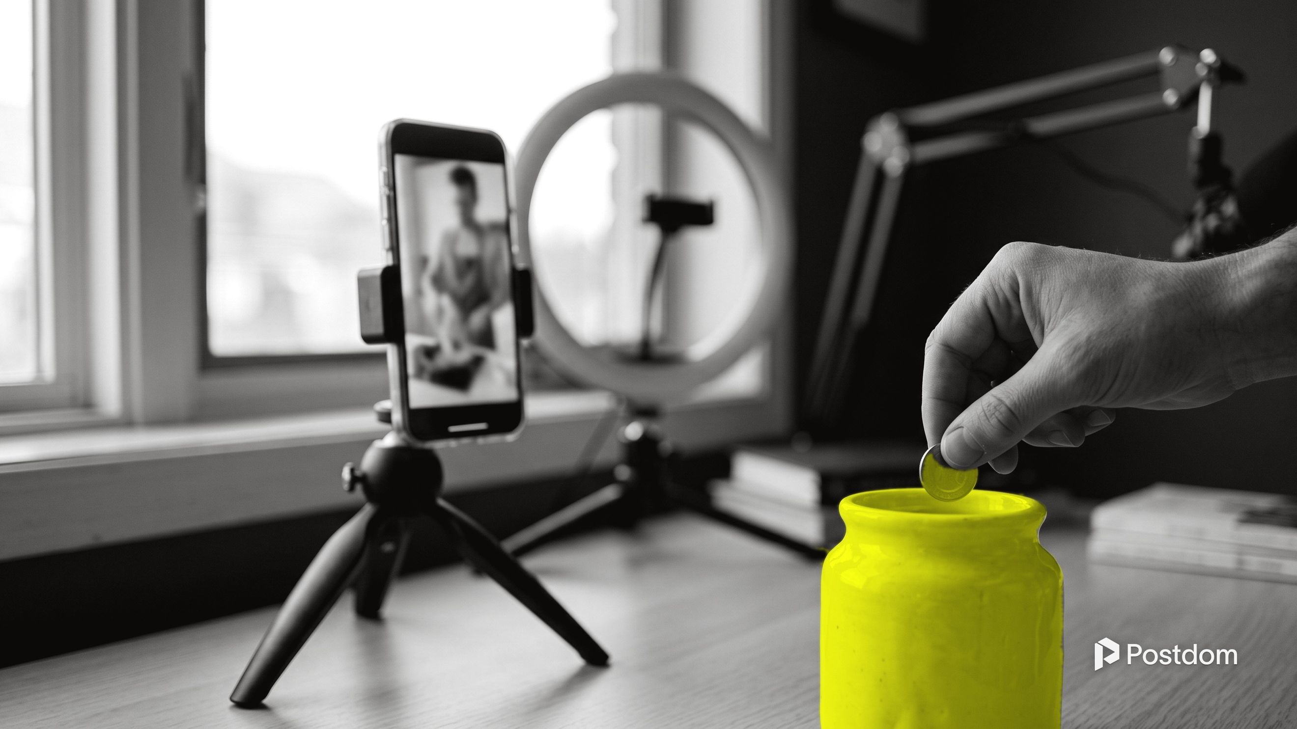A phone on a mini tripod films a creator's desk while a hand drops a coin into a yellow jar