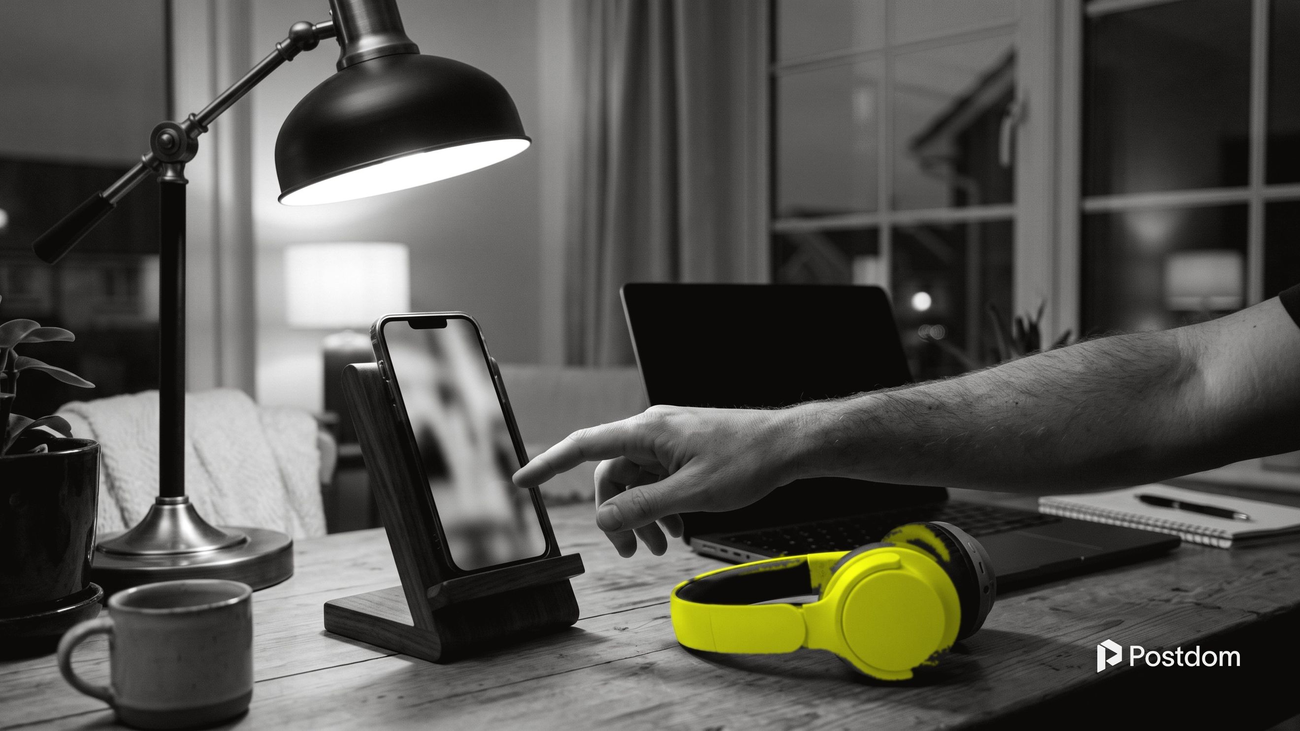How to schedule YouTube Shorts: a creator taps a phone standing upright in a desk stand next to yellow headphones and a laptop in the evening