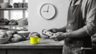 Best time to post on Facebook: a baker in a linen apron holds a phone over fresh loaves at the bakery counter, a yellow enamel mug beside him and a wall clock at nine behind