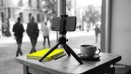 A phone on a small tripod beside a coffee cup and a yellow notebook at a cafe window, with people walking past outside