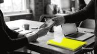 Social media approval workflow: one colleague hands a phone to another across a shared table to review a post, with a laptop and a yellow folder