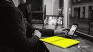 A cafe owner after closing works at a laptop with a phone in a stand beside it, both playing a street clip, next to a yellow notebook