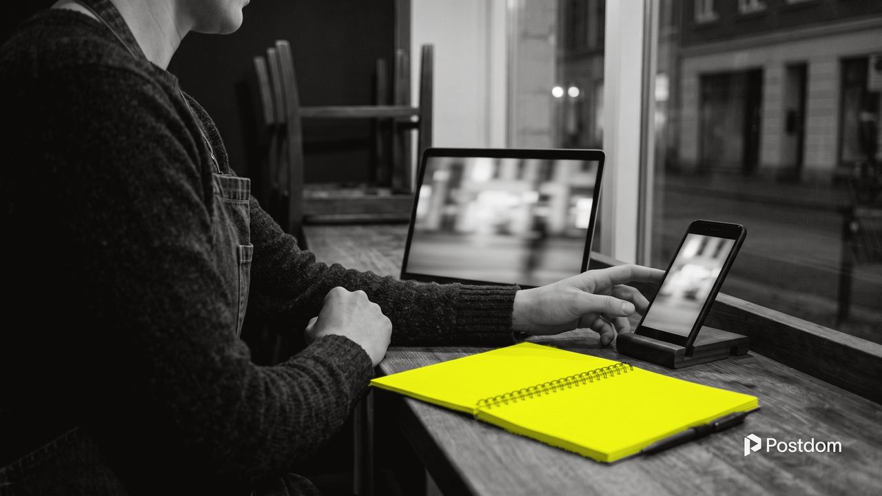 A cafe owner after closing, at a laptop with a phone in a stand beside it, both screens playing a blurred street clip, a yellow notebook on the counter