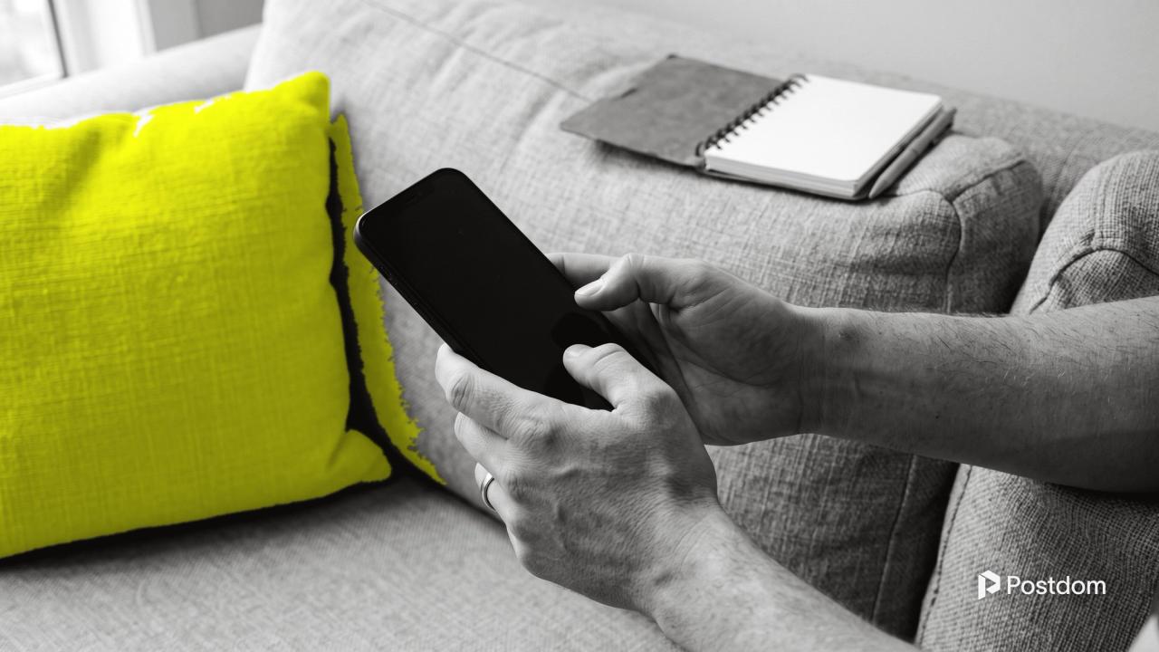 Hands scrolling a phone on a grey sofa beside a cushion, with an open notebook on the armrest