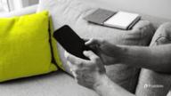 Hands scrolling TikTok on a phone on a grey sofa beside a yellow cushion, with an open notebook on the armrest