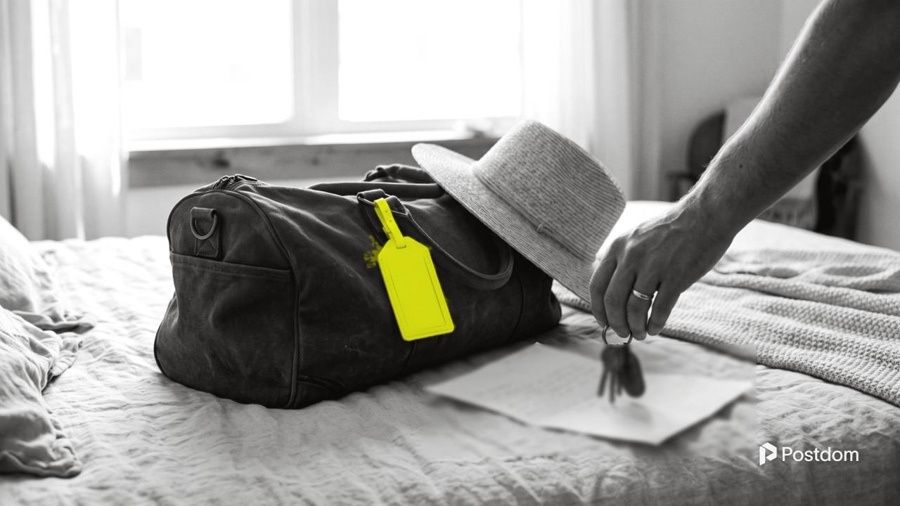 A packed travel bag with a yellow luggage tag and a sun hat on a bed, as a hand sets keys down on a handwritten note