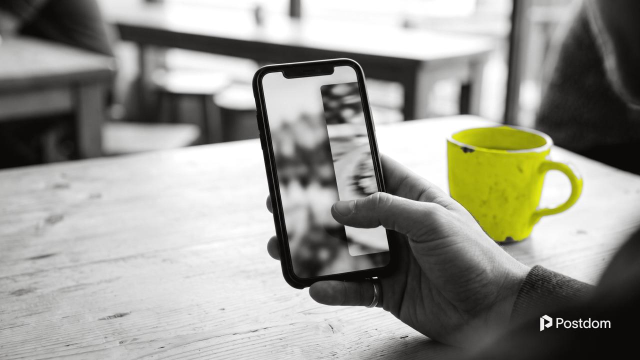 A hand holding a phone at a cafe table, thumb mid-swipe as one photo slides into the next, with a yellow mug beside it