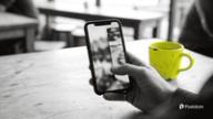 A hand holds a phone at a cafe table, thumb mid-swipe through a TikTok slideshow made in photo mode, with a yellow mug beside it