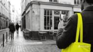 A shop owner with a yellow tote bag films a corner cafe on a cobbled street with a phone, a real place to open a TikTok on