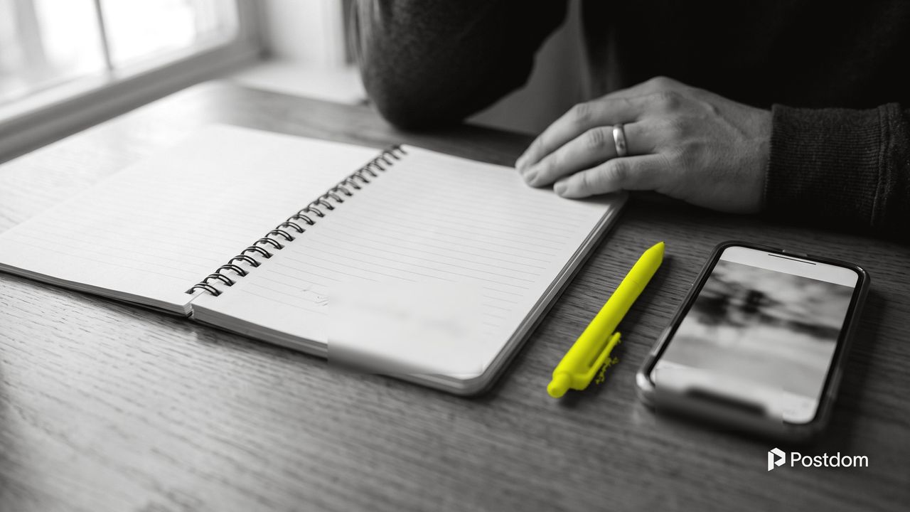 An open notebook with a pen set down beside it and a phone on a wooden table, a yellow pen and a hand resting nearby