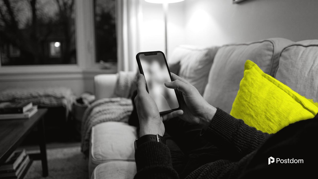 Best time to post on YouTube Shorts: a person on a sofa holds a phone in the evening next to a yellow cushion, dusk outside the window