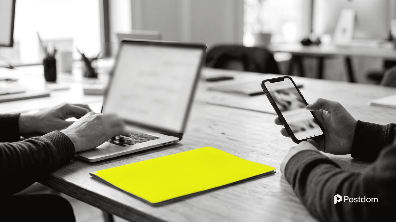 Social media management software: two colleagues at a shared table, one working on a content calendar on a laptop, the other holding a phone, a yellow folder between them