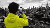 A person in a yellow jacket, seen from behind on a rooftop, films the city skyline on a phone for Snapchat