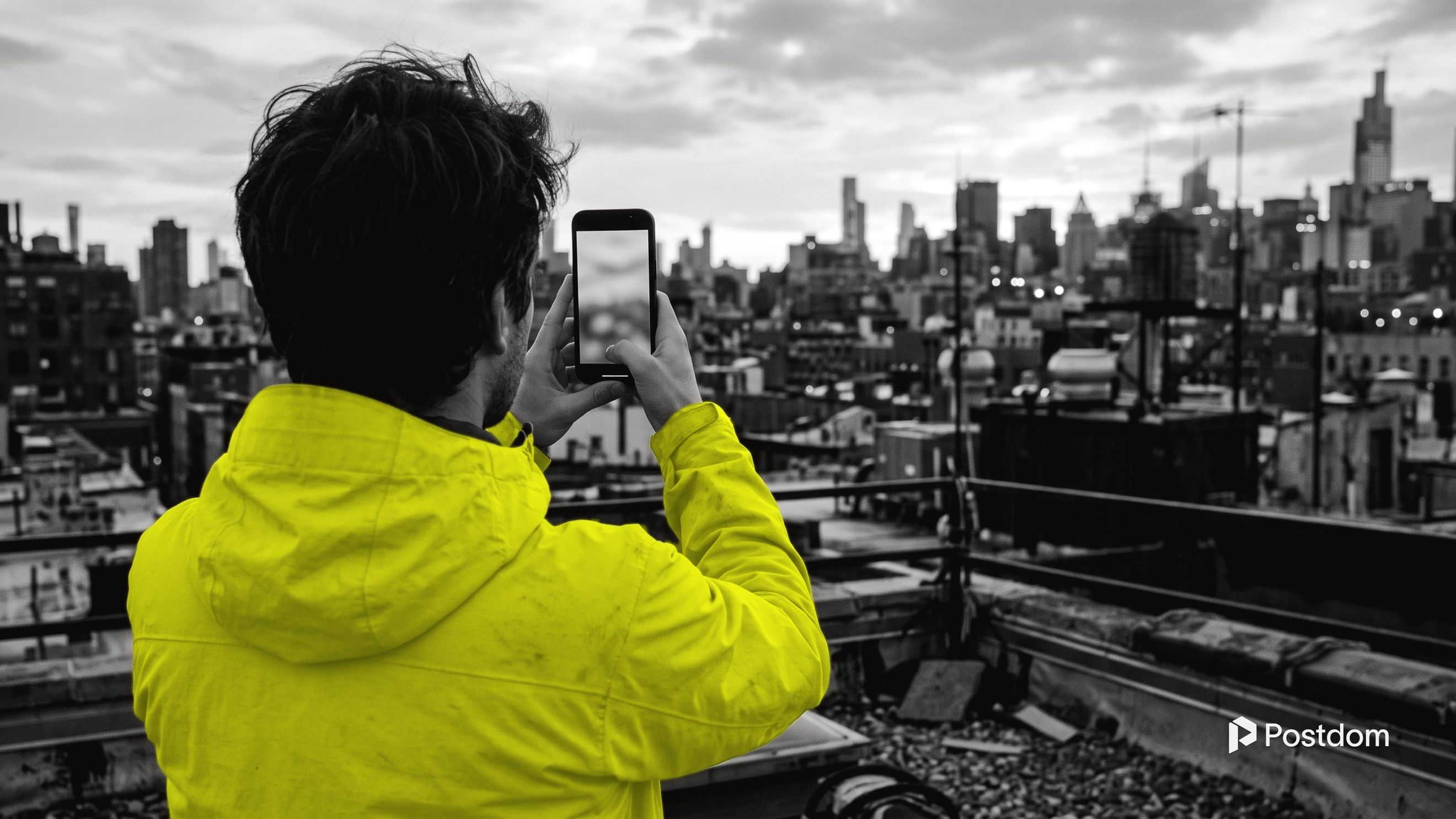 A person in a yellow jacket, seen from behind on a rooftop, filming the city skyline on a phone