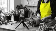A phone on a mini tripod films a florist in a yellow apron wrapping a bouquet at the shop counter for an Instagram Reel