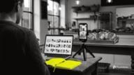 A freelancer at a bakery table works on a laptop while a phone on a small tripod films a tray of bread, a yellow notebook beside it