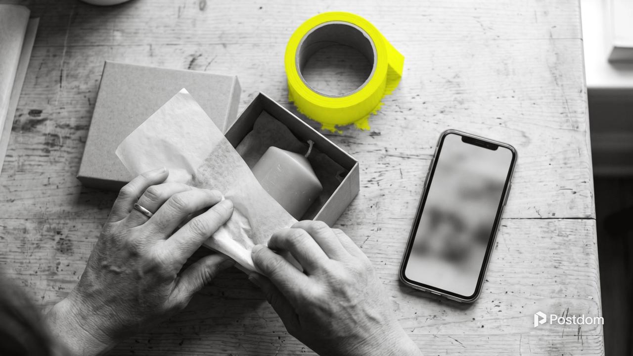 Hands wrap a handmade candle in tissue paper and pack it into a kraft box beside a phone.