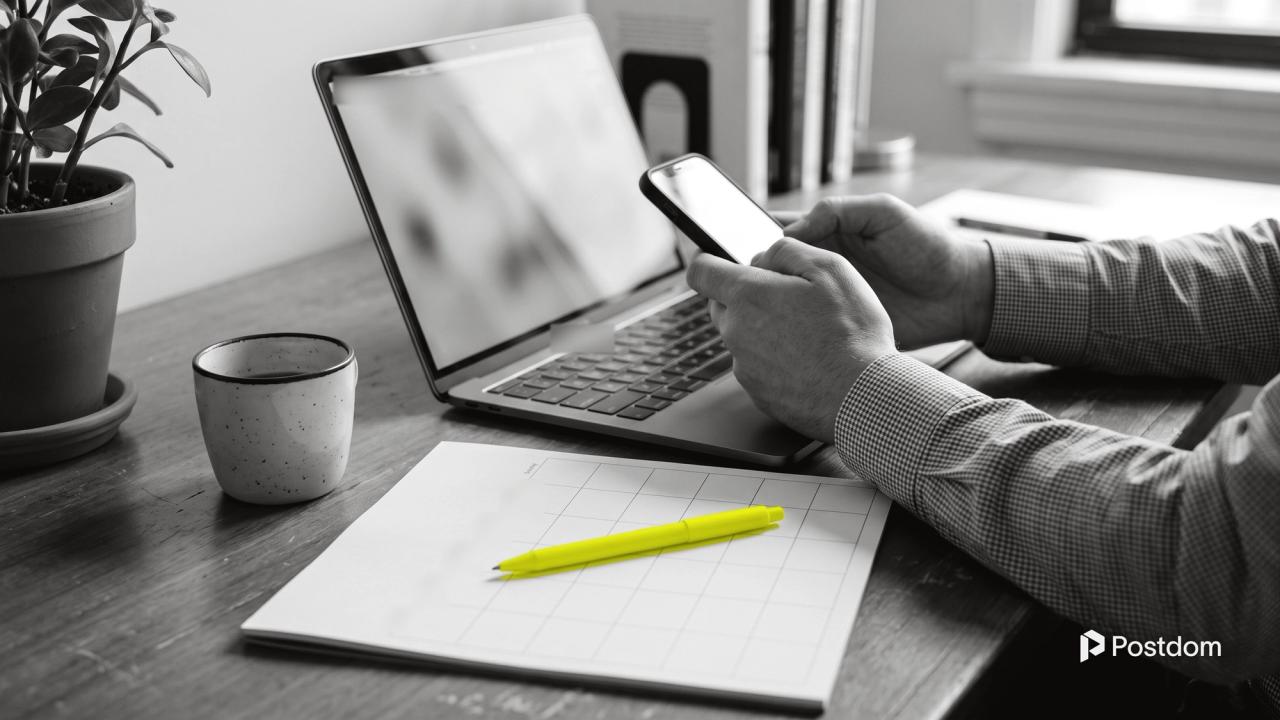 How to schedule LinkedIn posts: a professional holds a phone over a laptop next to a blank desk calendar and a yellow pen