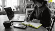 A creator at a kitchen table writes TikTok earnings figures in a yellow notebook beside a laptop, a phone and a cup of coffee