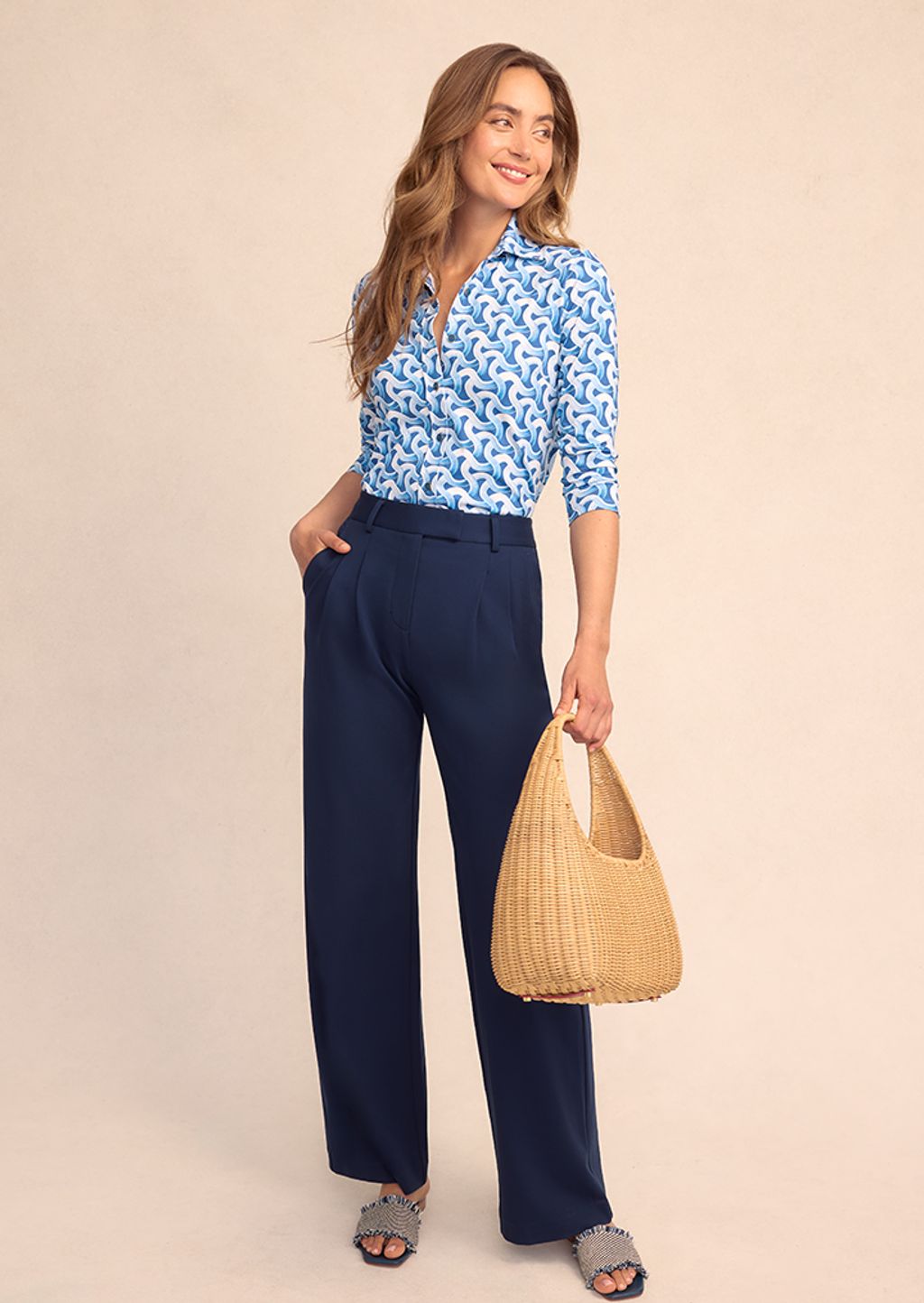 Model wearing navy slacks and blue on blue printed top with a wicker handbag.