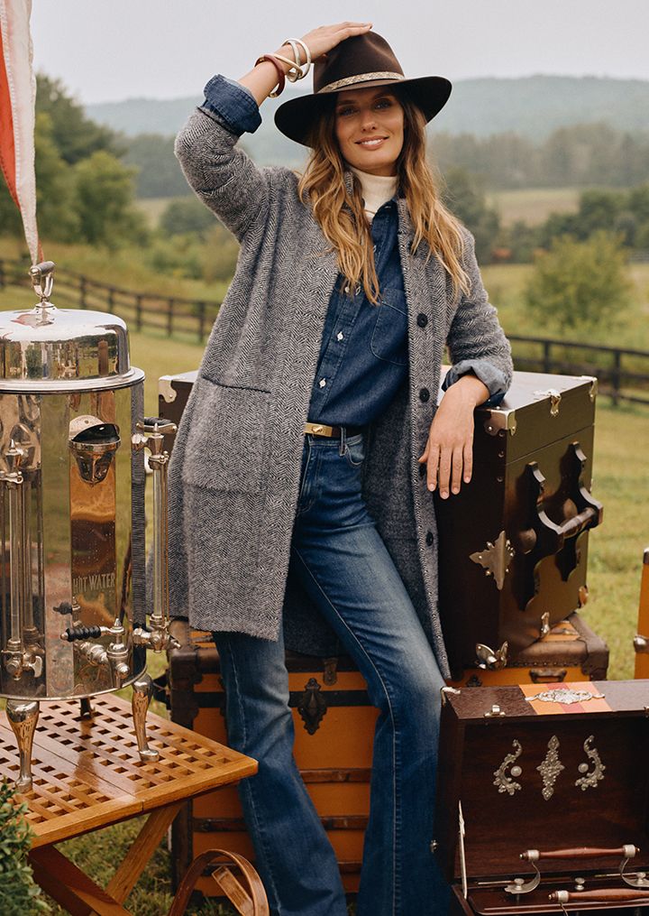 woman standing against steamer trunks outside wearing jeans, a denim shirt and a gray herringbone cardi coat