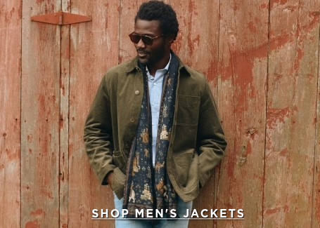 man leaning against a barn door wearing an olive suede jacket, jeans and a printed scarf