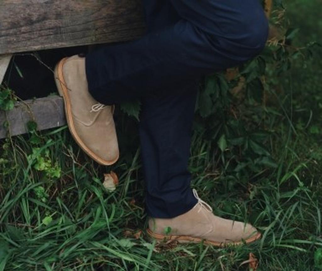 Legs in dark blue pants and beige chukka boots, one foot on a wooden structure in grass.