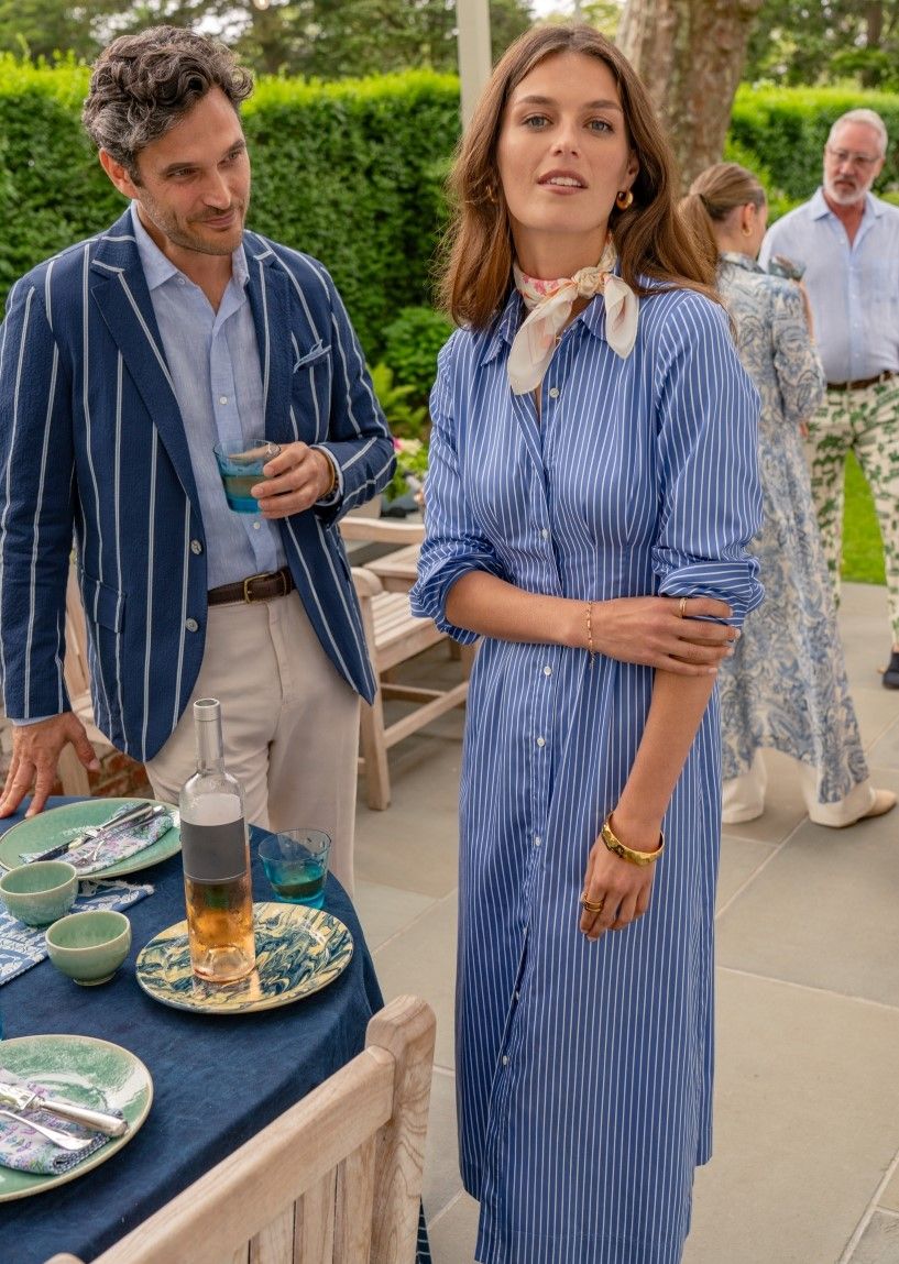 woman standing outside on a patio at a dinner party wearing a blue midi shirtdress with white hairline stripe and silk scarf tied at neck