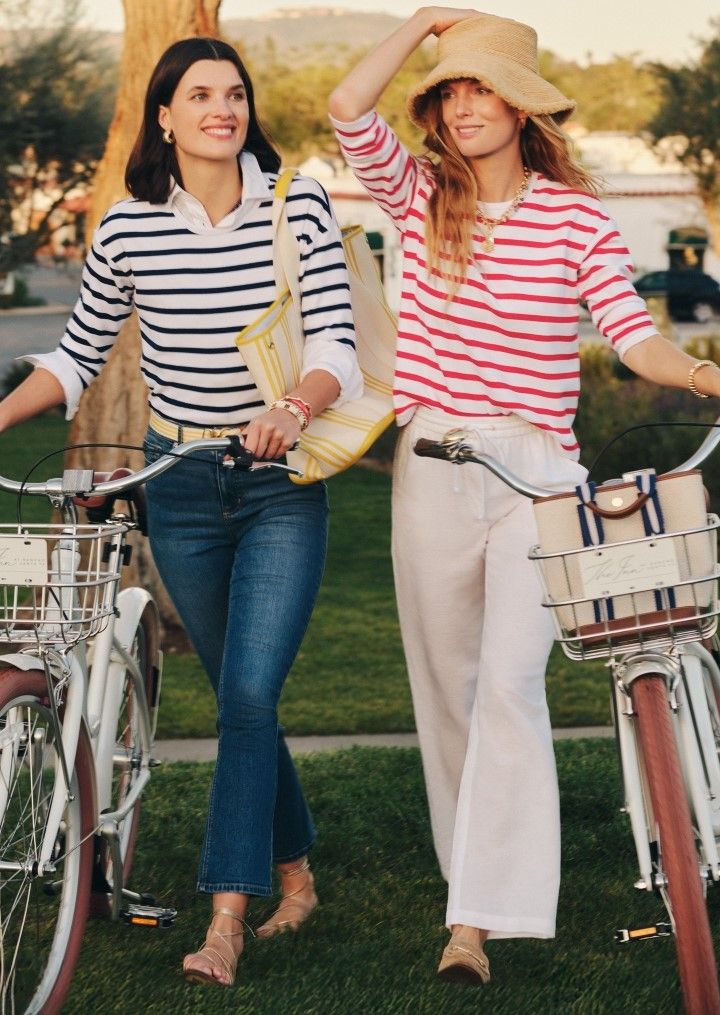 Two smiling women walk alongside their bicycles on a sunny day; one in a navy striped top and jeans, the other in a red striped top, white pants, and a straw hat.