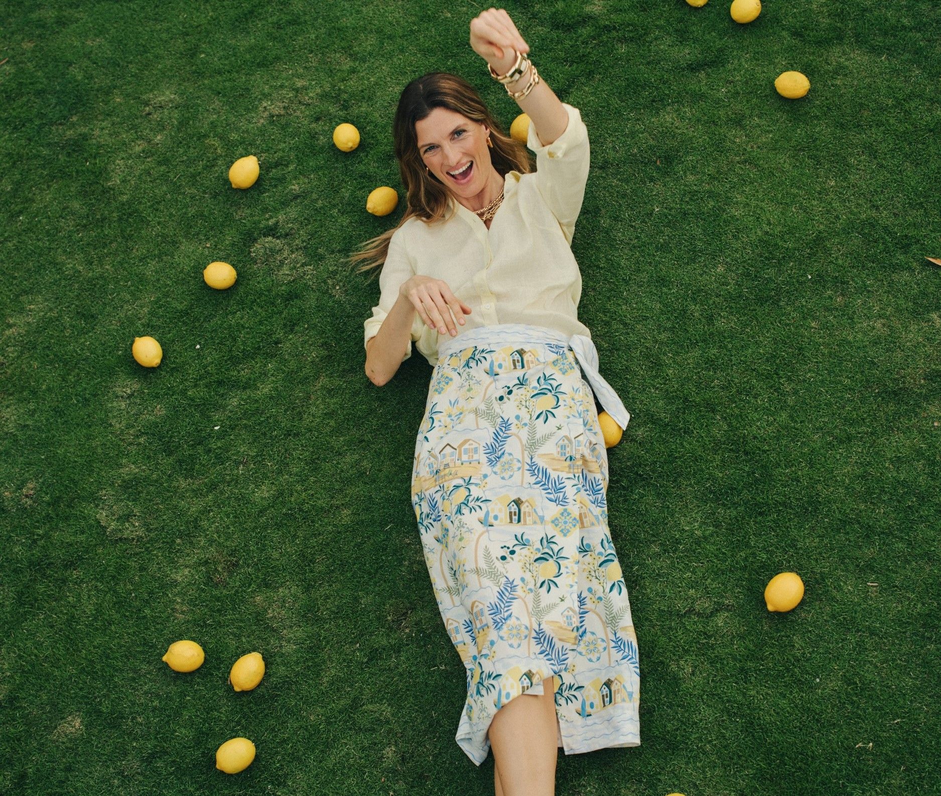 A smiling woman in a yellow shirt and patterned skirt lies on green grass, surrounded by lemons, with one arm raised.