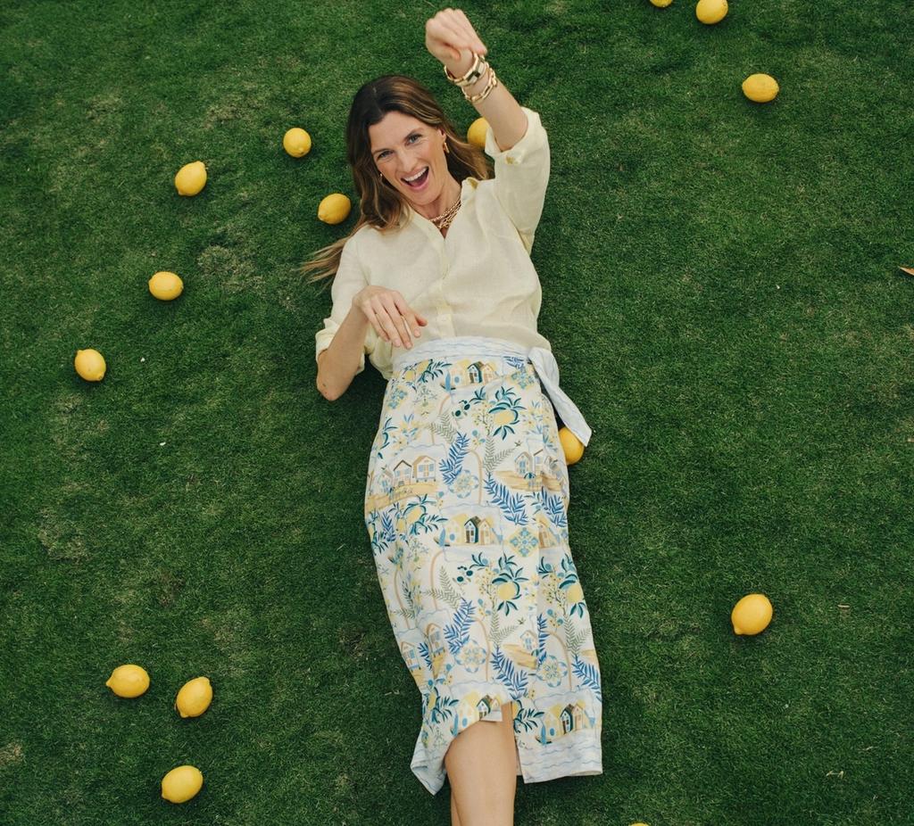 A smiling woman in a yellow shirt and patterned skirt lies on green grass, surrounded by lemons, with one arm raised.