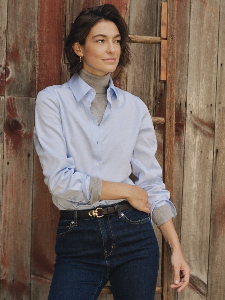 A woman with short dark hair wears a blue shirt over a grey turtleneck and jeans, leaning on a weathered wooden wall.