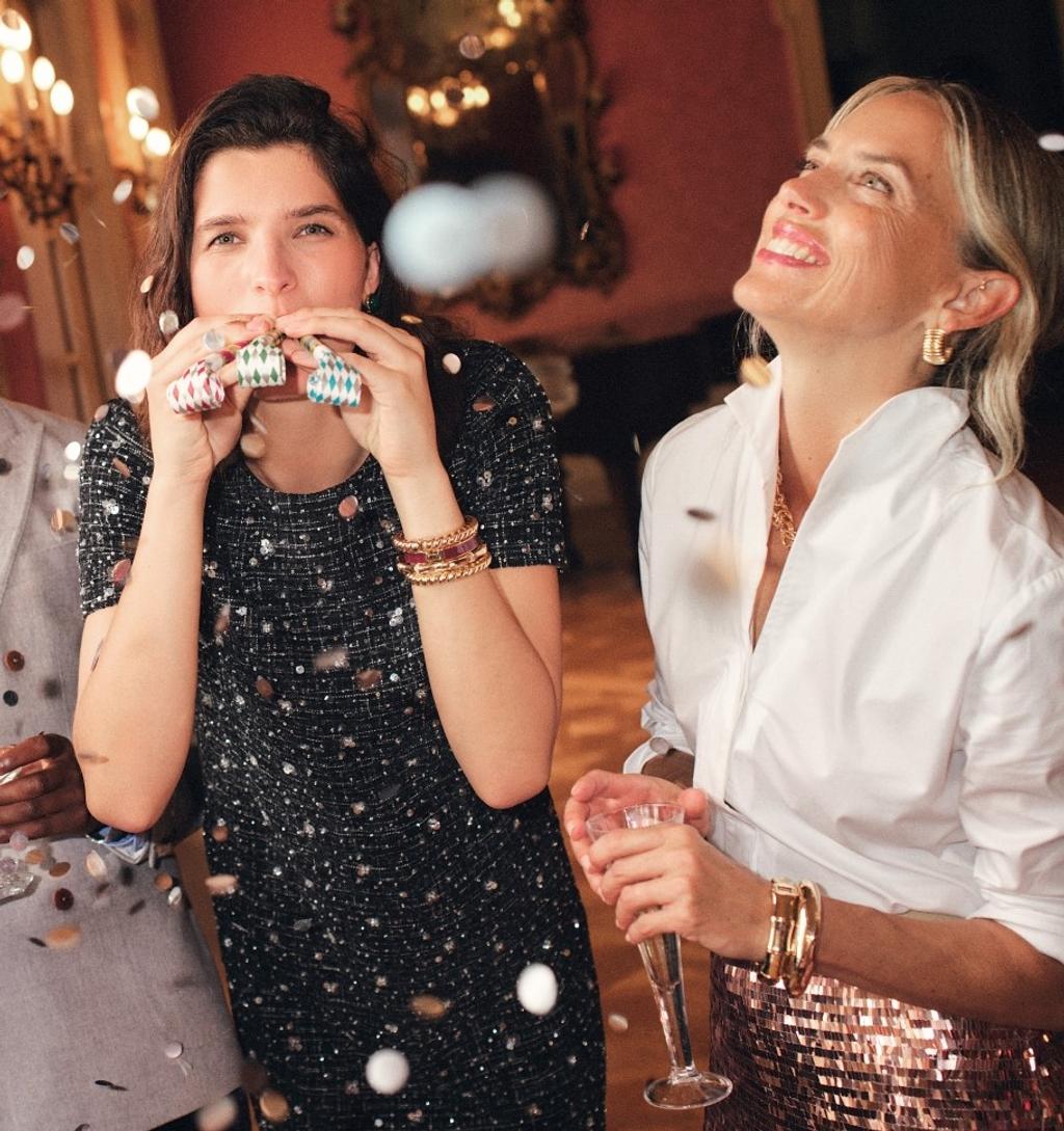 Two women at a party with falling confetti; one in a sparkly dress blows party favors, the other in a sequined skirt smiles with a champagne flute.