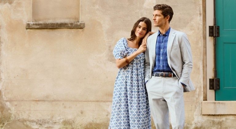 man and woman standing outside in front of stone house. Woman wearing white puff sleeve flowy dress with blue geo pattern. Man wearing stone slacks with matching blazer and blue tonal plaid shirt