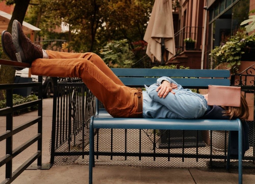 man laying on bench in chambray shirt and camel corduroy pants with a leather backed book over his eyes