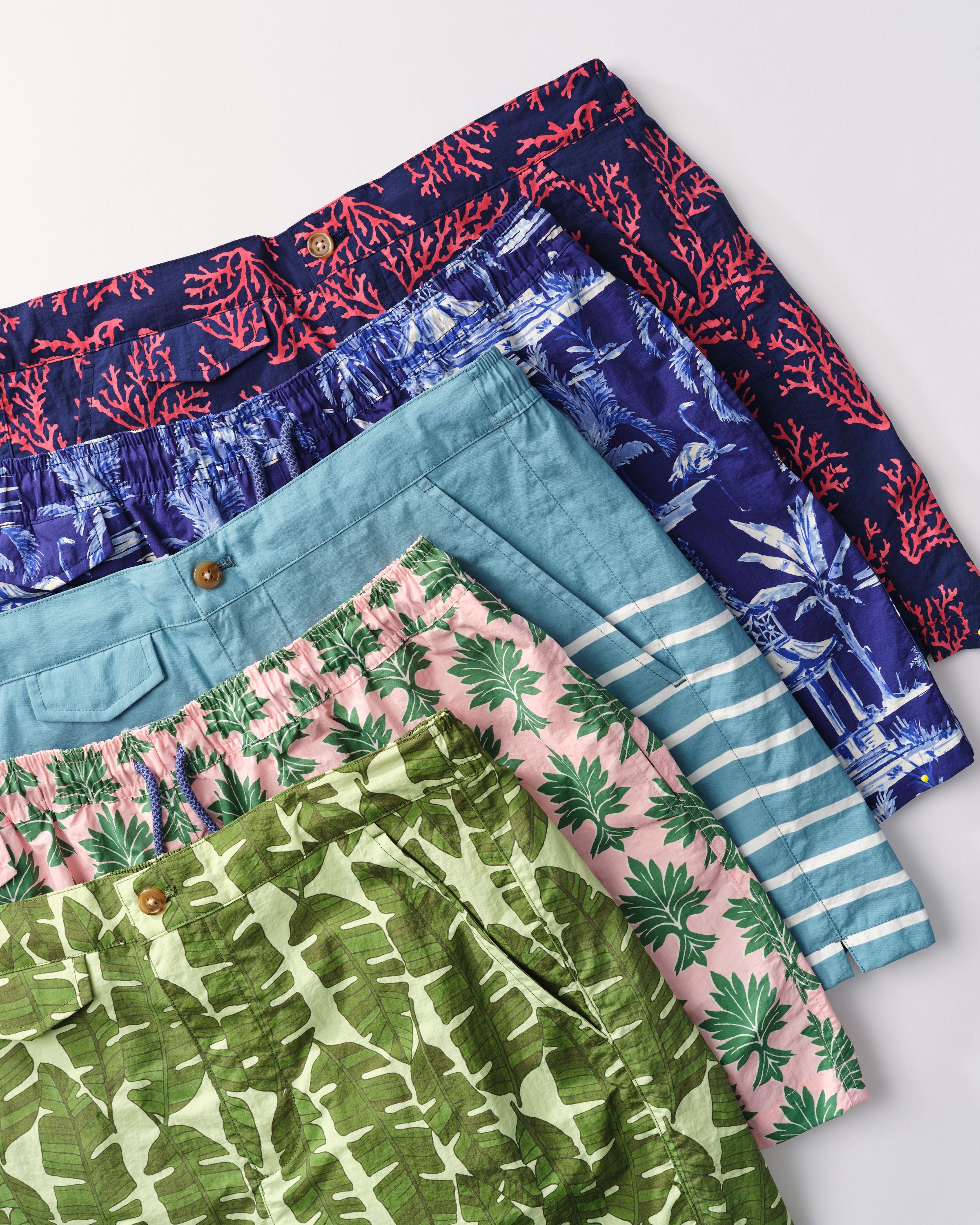 A diagonal stack of five colorful shorts with various patterns, including coral, tropical scenes, stripes, and leaf designs.
