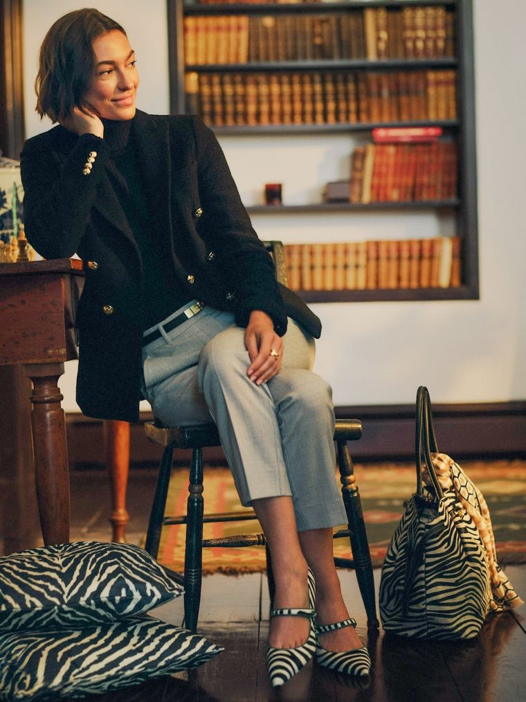 A smiling woman in a black blazer, grey pants, and zebra-print shoes sits in a study with bookshelves and zebra-print decor.